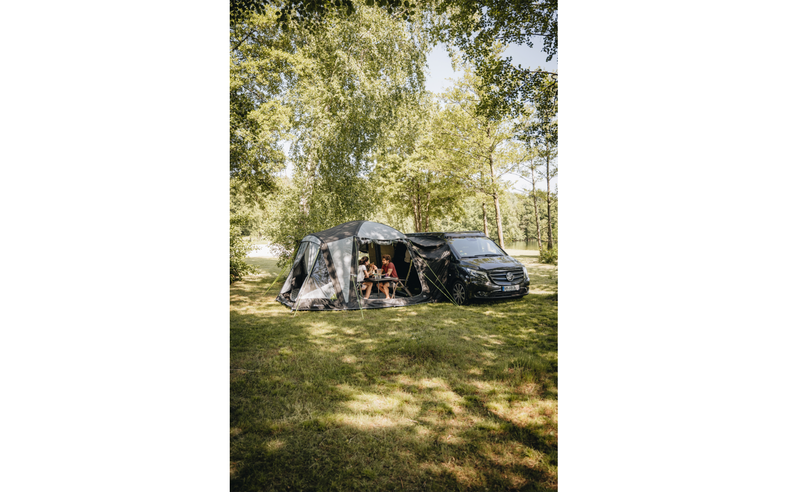 Black camper van with attached gray awning tent; two people sitting at a small table inside the awning; grass, tall trees and a body of water in the background; sunlight and shadows