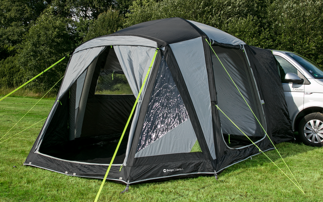 Ein graues Vorzelt ist an ein weißes Wohnmobil auf einer grünen Wiese angebracht, im Hintergrund sind Bäume und ein blauer Himmel mit Wolken sichtbar
