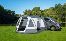Tent pitched on grass next to a gray van, with trees and blue sky in the background