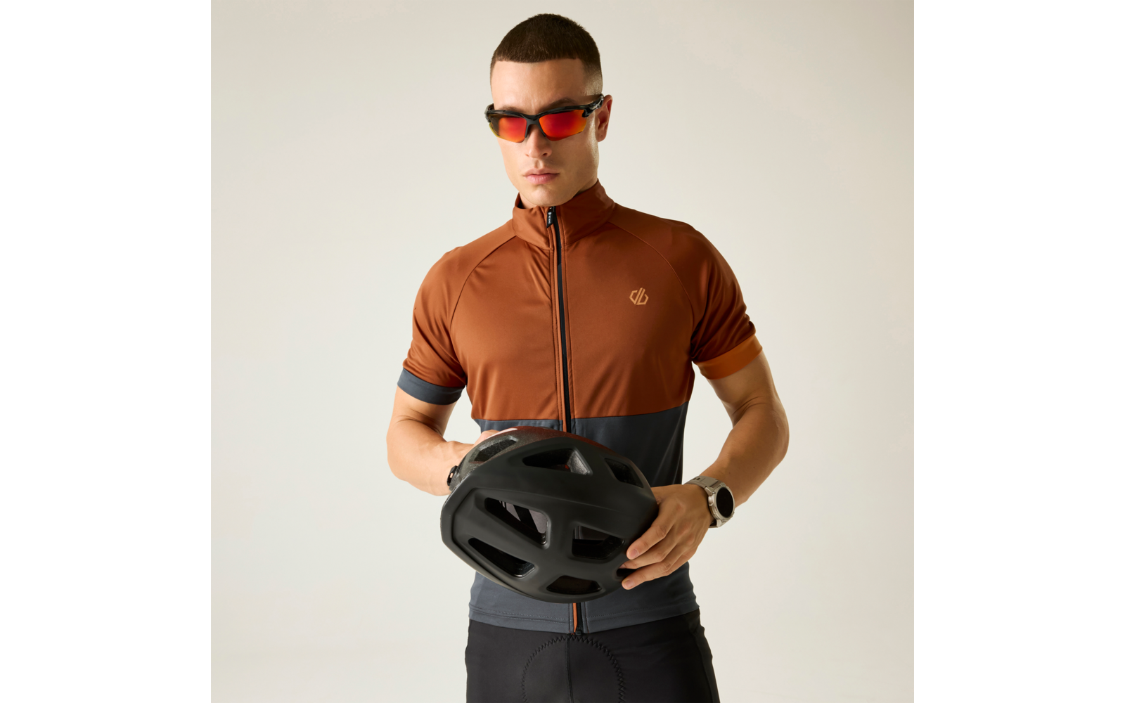 Hombre con maillot de ciclismo marrón y gris oscuro y culotte negro, con gafas de sol rojas, sosteniendo un casco de bicicleta negro frente al torso sobre fondo claro