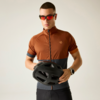 Hombre con maillot de ciclismo marrón y gris oscuro y culotte negro, con gafas de sol rojas, sosteniendo un casco de bicicleta negro frente al torso sobre fondo claro