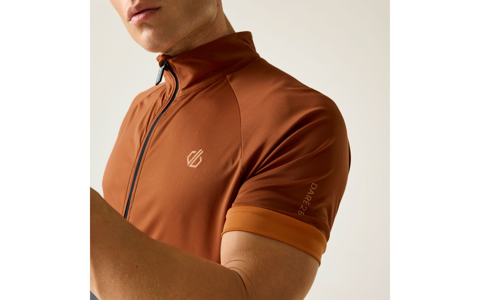 Parte superior del cuerpo de una persona, barbilla y boca recortadas visibles, prenda marrón ajustada de manga corta con cremallera visible, pequeño logo en el pecho y texto en la manga