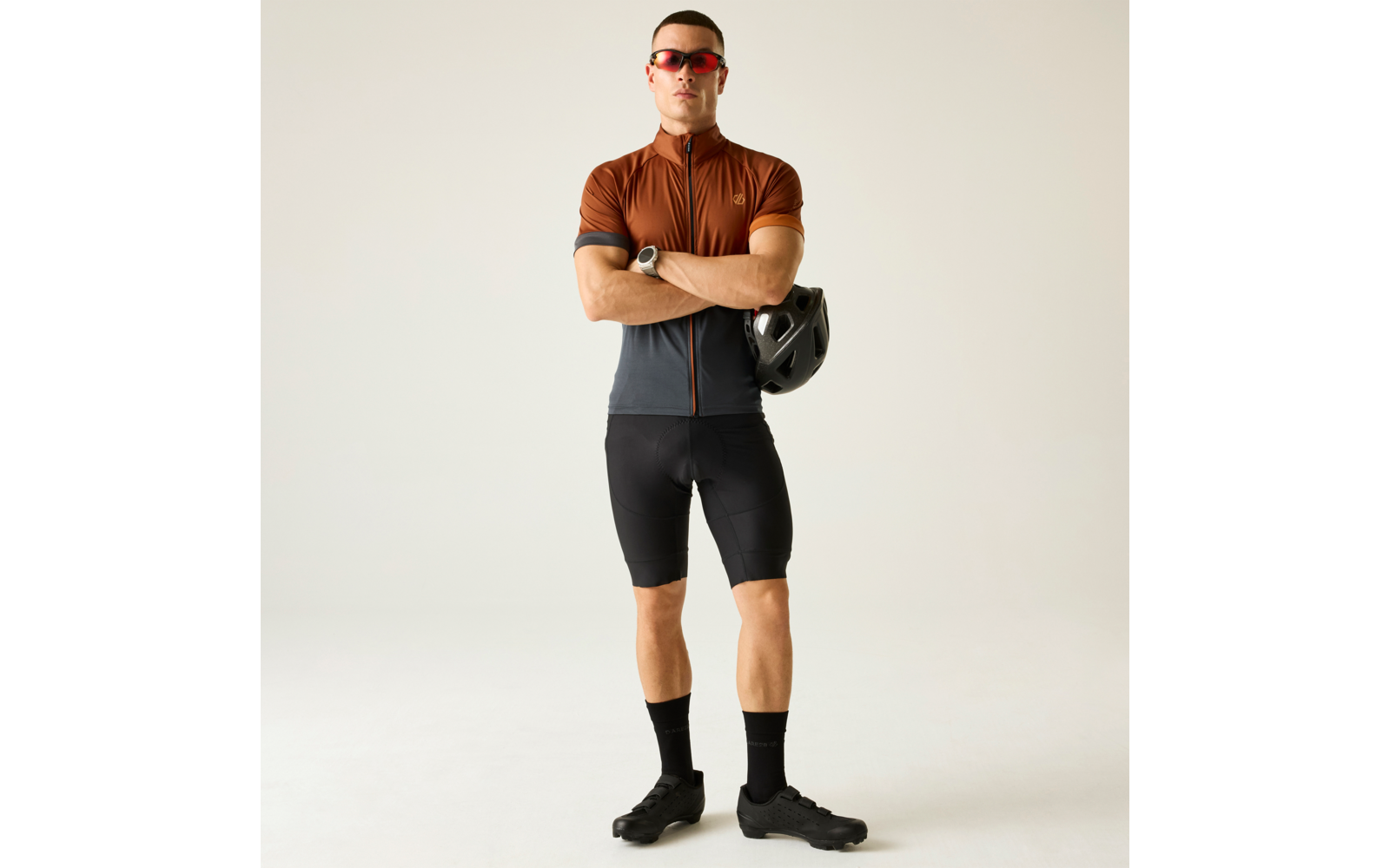 Persona de pie con los brazos cruzados, sostiene un casco de bicicleta negro, lleva un maillot de ciclismo marrón-gris, pantalones cortos negros, calcetines y zapatillas negras, gafas de sol rojas, reloj en la muñeca, fondo claro