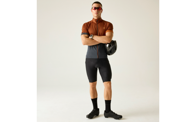 Persona de pie con los brazos cruzados, sostiene un casco de bicicleta negro, lleva un maillot de ciclismo marrón-gris, pantalones cortos negros, calcetines y zapatillas negras, gafas de sol rojas, reloj en la muñeca, fondo claro