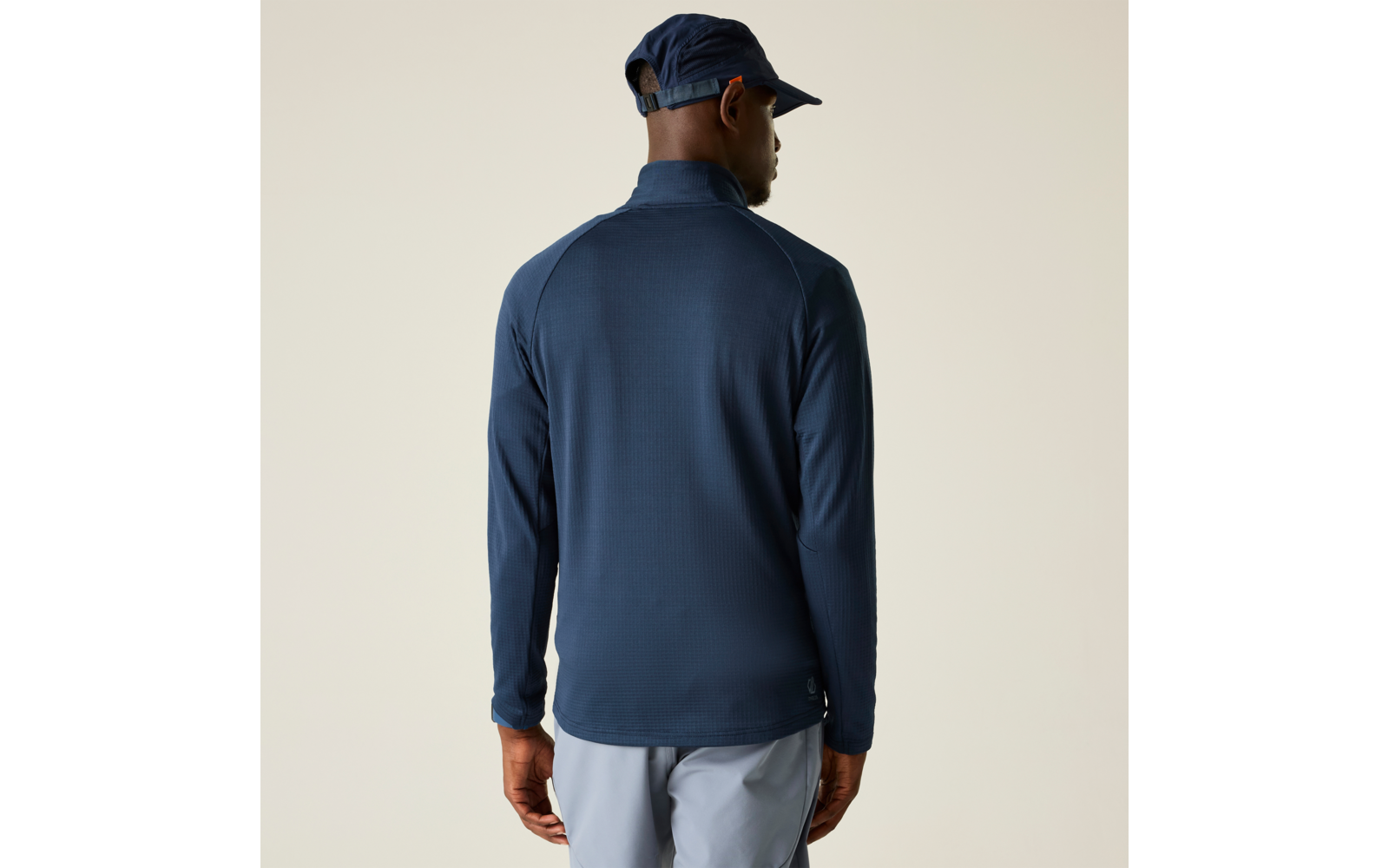 Person von hinten, trägt dunkelblaues langärmeliges Oberteil mit Stehkragen, dunkelblaue Kappe und hellgraue Hose vor beigem Hintergrund