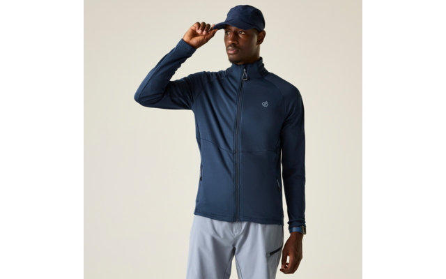 Person steht vor hellem Hintergrund, trägt dunkelblaue Reißverschlussjacke mit Stehkragen und kleinem Logo auf der Brust, dunkelblaue Cap, hellgraue Hosen, rechte Hand hebt die Capkrempe, linke Hand an der Seite mit Armbanduhr