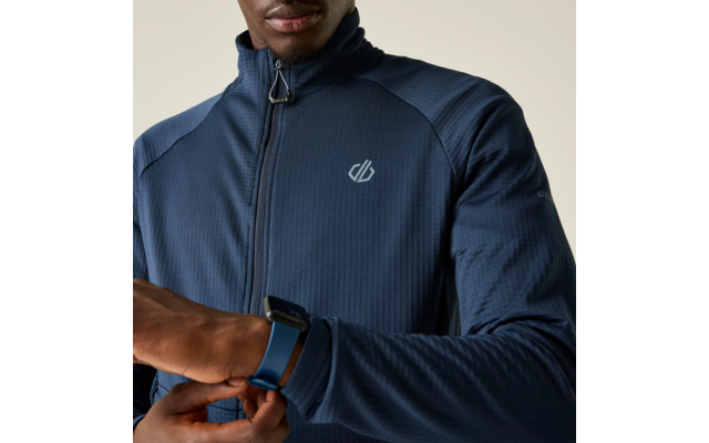 Oberkörper einer Person in dunkelblauer Jacke mit Reißverschluss und kleinem weißem Logo, justiert eine blaue Smartwatch am linken Handgelenk; Kinn und Teil des Gesichts sichtbar; heller beiger Hintergrund