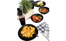 Teller mit Pasta, daneben eine Serviette und ein Glas Rotwein auf einem Holztisch, im Hintergrund Kräuter und Knoblauch