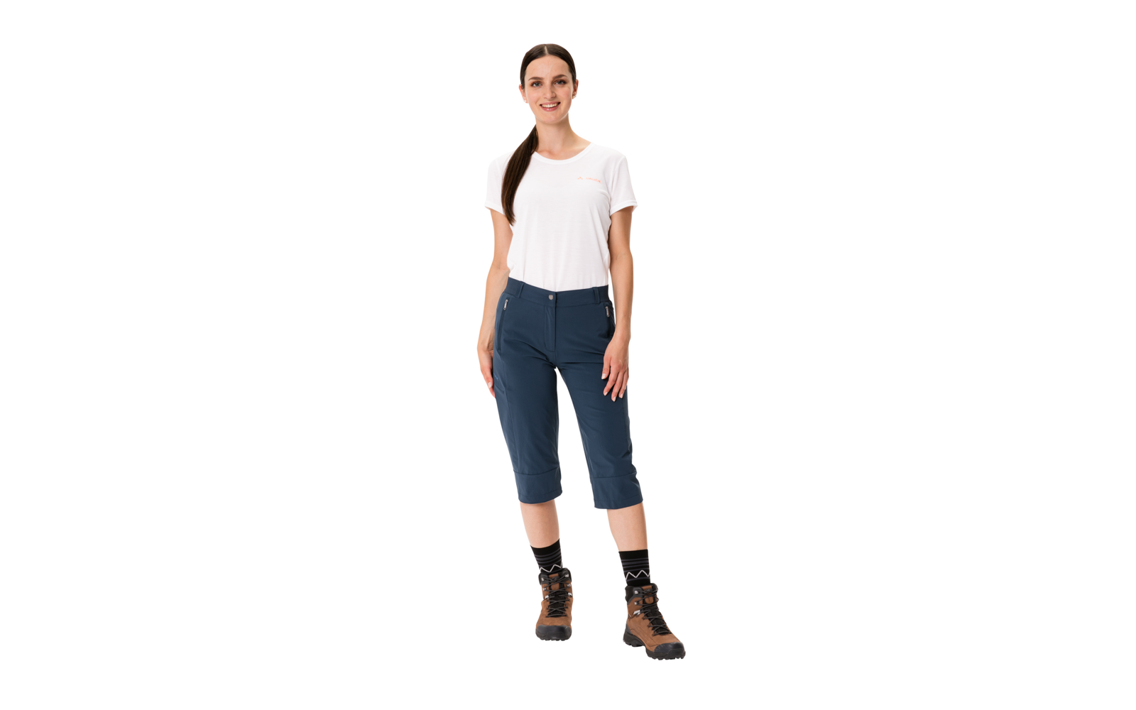 Woman standing facing forward, wearing a white T-shirt, navy knee-length pants, black socks and brown hiking boots, hair in a ponytail