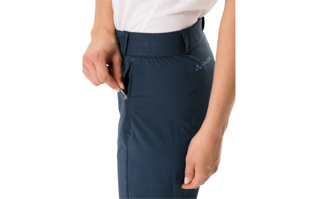 Waist to mid-thigh of a person wearing dark blue pants with elastic waistband and side zip pocket; left hand holding the zipper, white t-shirt visible at top edge