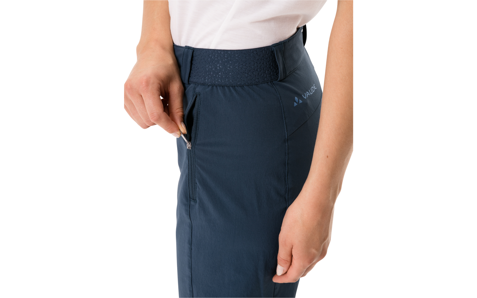 Waist to mid-thigh of a person wearing dark blue pants with elastic waistband and side zip pocket; left hand holding the zipper, white t-shirt visible at top edge