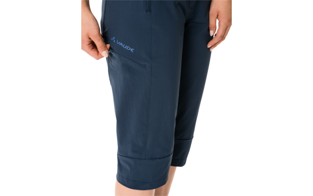 Close-up from hips to lower calves wearing dark blue knee-length pants with visible "VAUDE" logo, one hand touching a pocket, white background