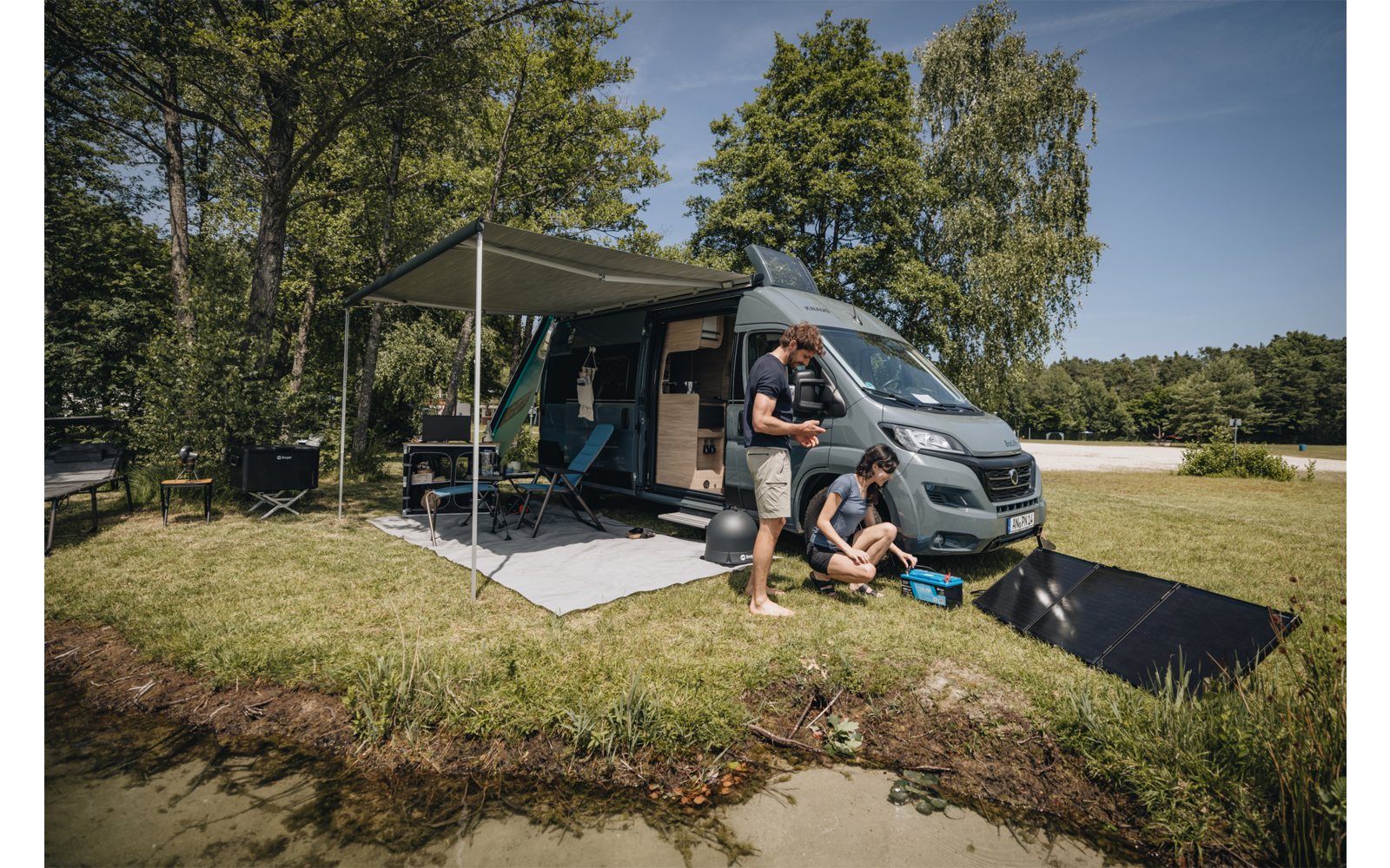 Zwei Personen neben einem geparkten Campingbus auf einer Wiese am Seeufer; ausgefahrene Markise, offene Schiebetür, Campingstühle und -tisch, Angelzubehör am Wasserrand, Bäume im Hintergrund