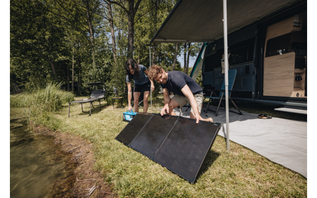 Zwei Personen neben einem Wohnmobil am See, sie stellen faltbare Solarpaneele auf der Wiese auf; ausgefahrene Markise, Bäume und Picknickbank im Hintergrund
