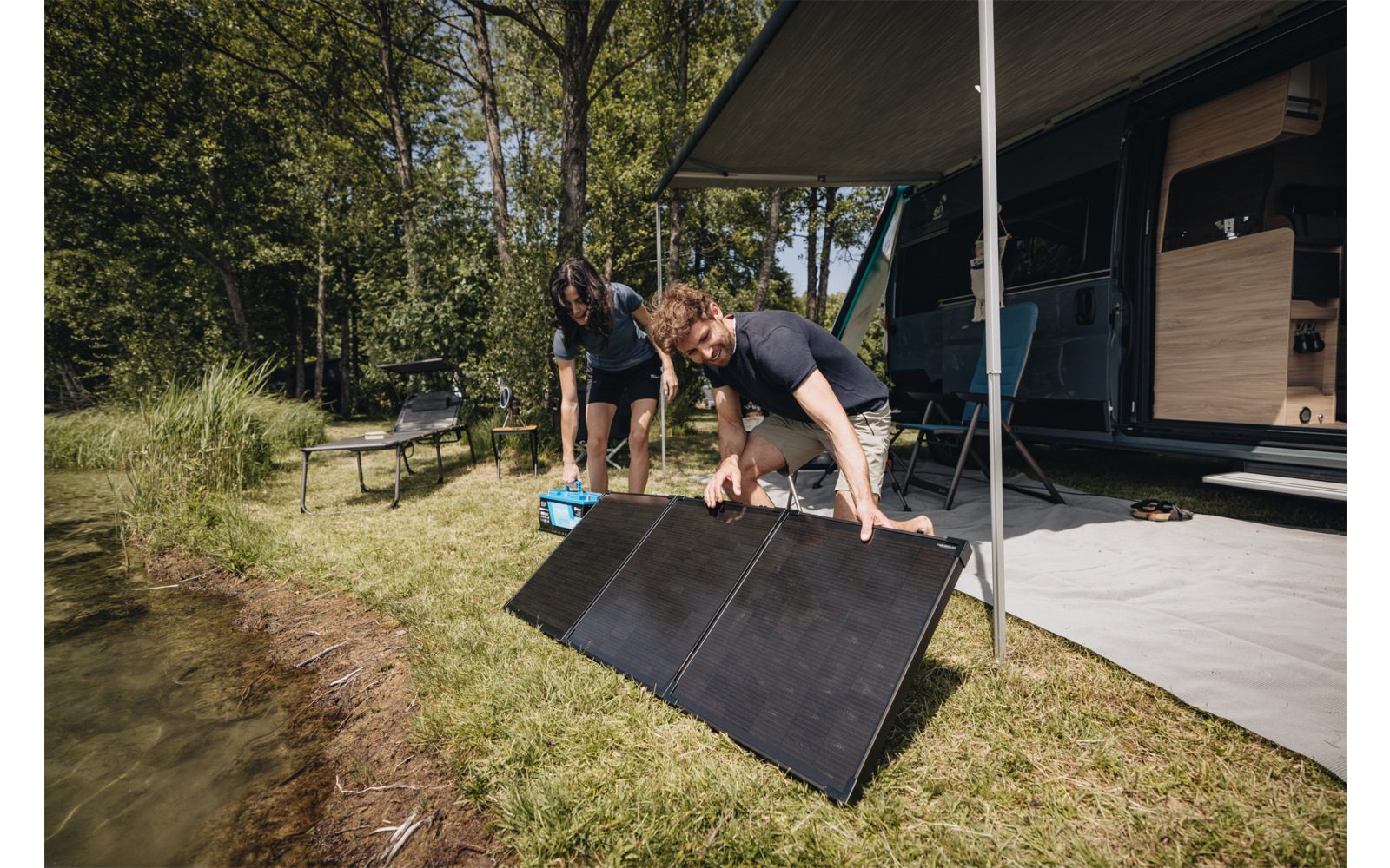 Zwei Personen neben einem Wohnmobil am See, sie stellen faltbare Solarpaneele auf der Wiese auf; ausgefahrene Markise, Bäume und Picknickbank im Hintergrund