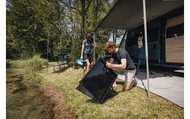 Zwei Personen neben einem Wohnwagen am See; ein Mann kniet und hält ein schwarzes rechteckiges Paneel, eine Frau trägt eine blaue Box; ausgefahrene Markise, Gras und Bäume