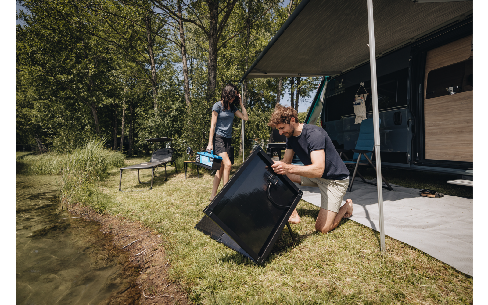 Zwei Personen neben einem Wohnwagen am See; ein Mann kniet und hält ein schwarzes rechteckiges Paneel, eine Frau trägt eine blaue Box; ausgefahrene Markise, Gras und Bäume