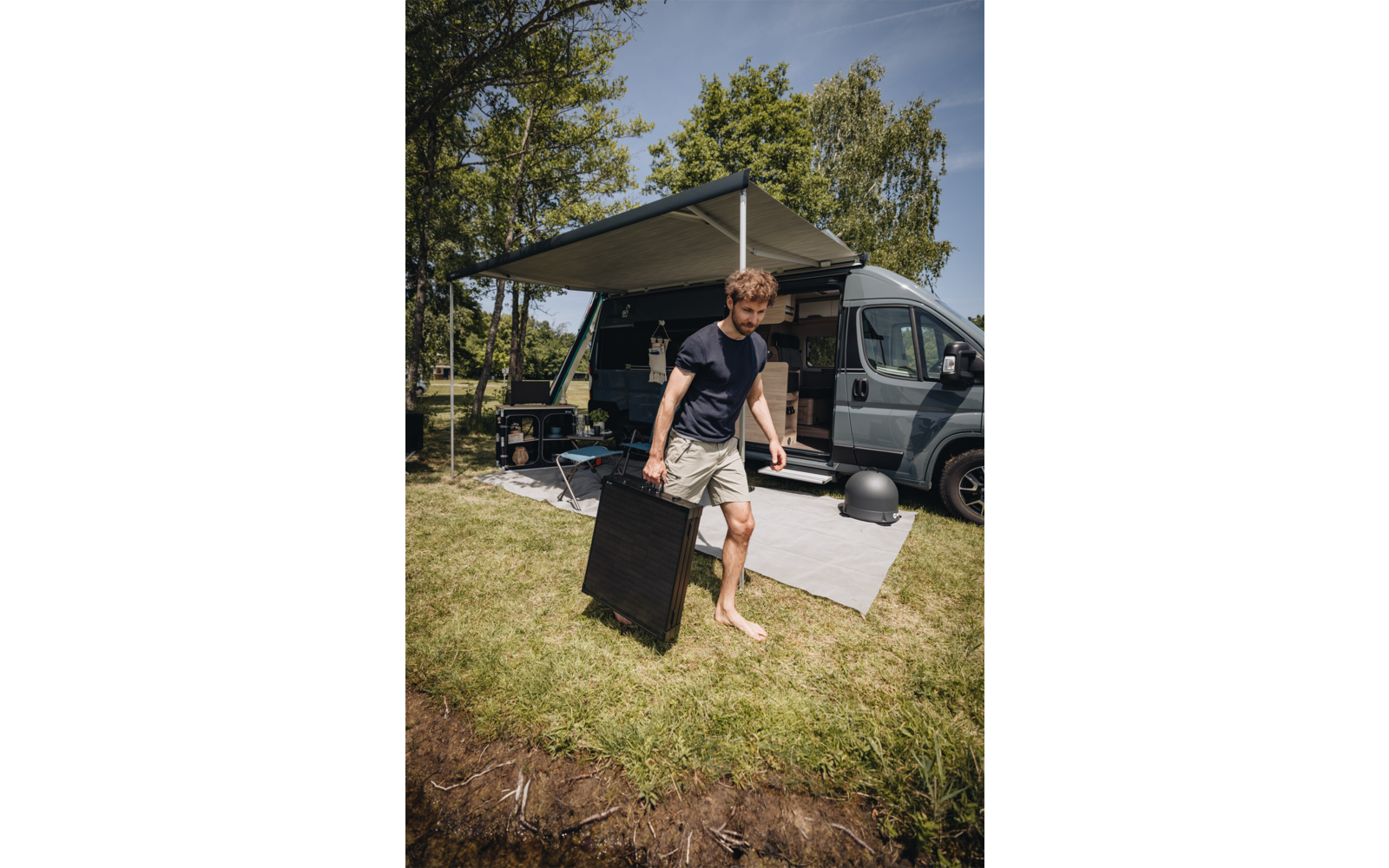 Mann ohne Schuhe trägt eine schwarze klappbare Platte vor einem geparkten Wohnmobil mit ausgefahrener Markise auf einer Grasfläche neben Wasser
