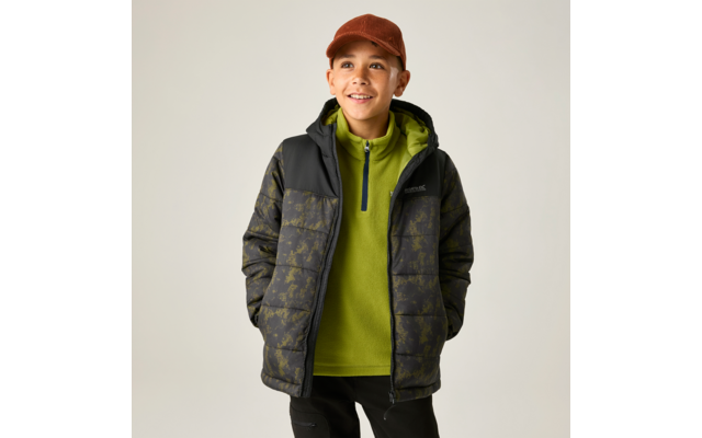 Junge vor hellem Hintergrund, braune Kappe, grüne Fleecejacke mit Reißverschluss, dunkle gesteppte Jacke mit Muster, Hände in den Taschen, Blick nach oben rechts