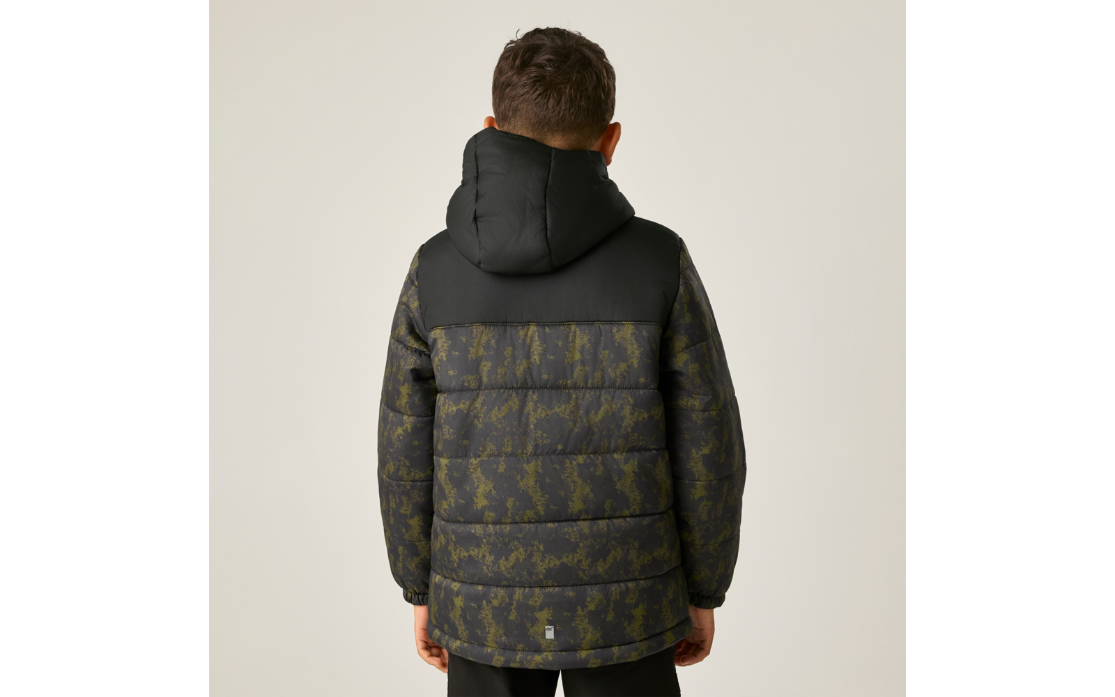 Person von hinten, trägt eine gesteppte Kapuzenjacke mit dunkelgrünem camouflageähnlichem Muster, schwarze Kapuze, kurze dunkle Haare, steht vor hellem einfarbigem Hintergrund