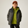Junge vor hellem Hintergrund, braune Kappe, grüne Fleecejacke mit Reißverschluss, dunkle gesteppte Jacke mit Muster, Hände in den Taschen, Blick nach oben rechts