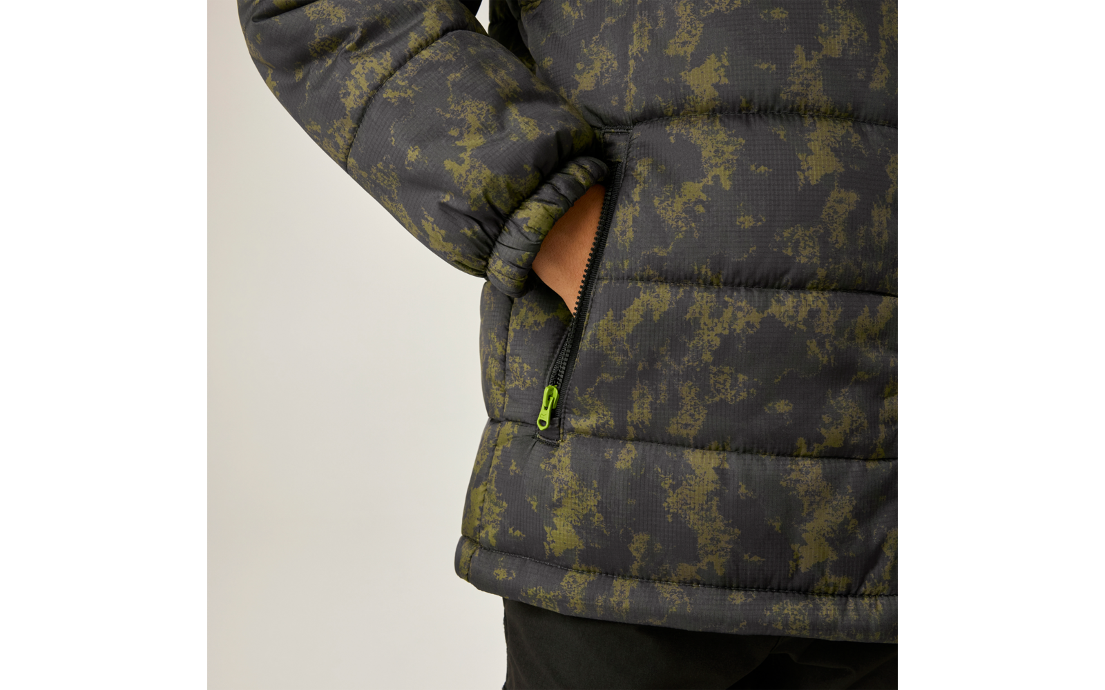 Nahaufnahme einer gesteppte Jacke mit grün-grauem Muster, Hand steckt in seitlicher Reißverschlusstasche mit neongrünem Zipper; unterer Jackenbereich und Teil einer schwarzen Hose sichtbar