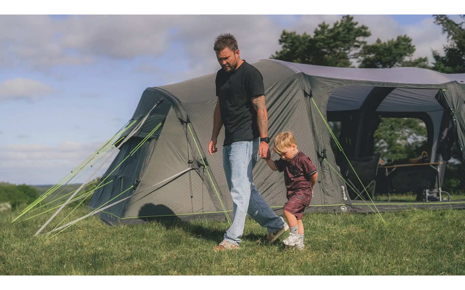 Mann und Kind neben einem grünen Zelt auf einer Wiese unter blauem Himmel