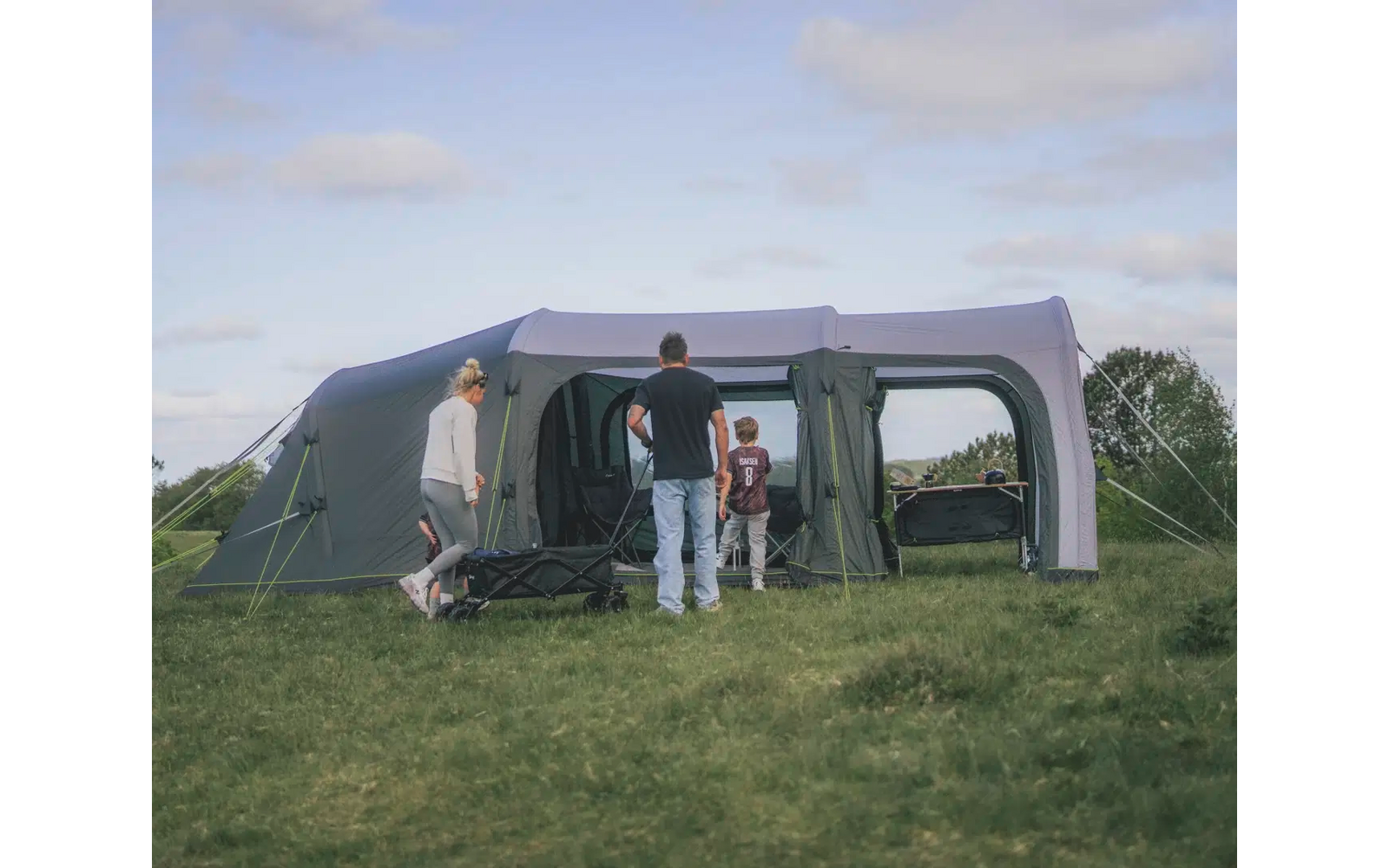 Zwei Personen stehen vor einem großen, grauen Zelt auf einem Campingplatz unter einem bewölkten Himmel