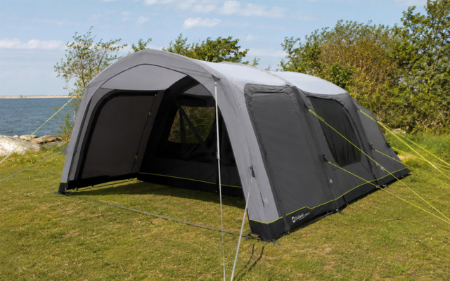 Grote grijze tent met open ingang op gras, scheerlijnen zichtbaar, water, bomen en bewolkte lucht op de achtergrond