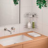 Lavabo rectangulaire blanc encastré dans un plan vasque en bois, robinet mural chromé et commande, étagère d'angle métallique à deux niveaux avec un vase blanc contenant des bâtonnets et une bouteille verte, serviette blanche pliée, miroir partiel et plantes vertes suspendues sur mur carrelé
