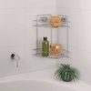 Étagère d'angle métallique sur mur carrelé de la salle de bain au-dessus de la baignoire ; étagère supérieure avec éponge ronde et objet plat, étagère inférieure avec bouteille verte et récipient transparent contenant des billes orange ; sur le bord de la baignoire un pot noir avec plante verte ; poignée de robinet chromée à gauche