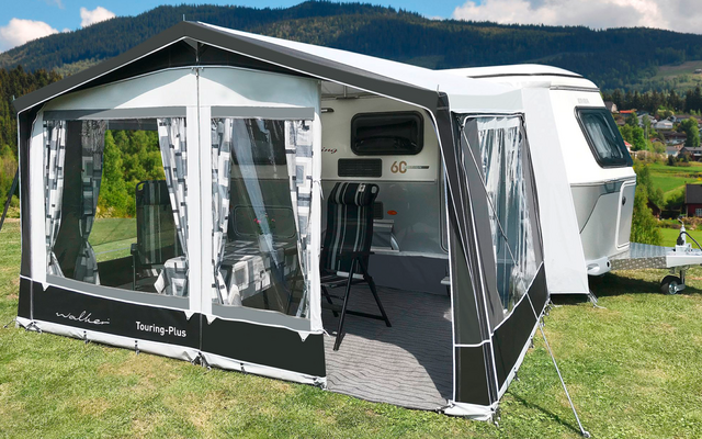 Wohnwagen mit angebautem Zelt auf einer Wiese, im Hintergrund Berge und Häuser