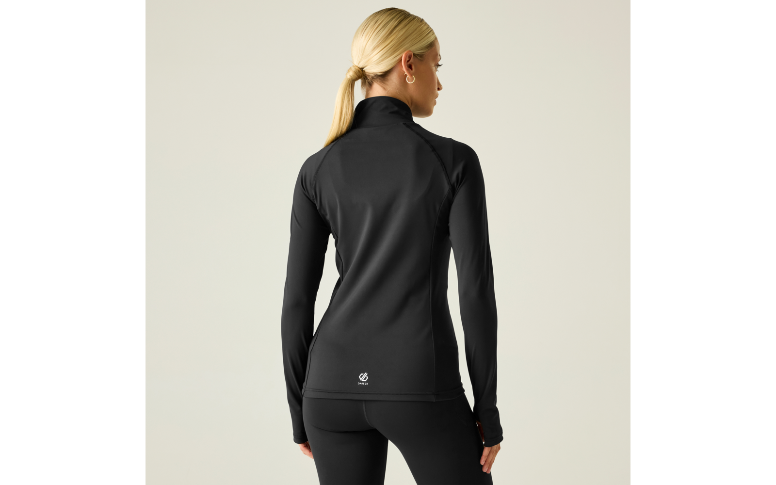 Mujer vista de espaldas, coleta rubia, chaqueta deportiva negra ajustada con cremallera y mallas negras, fondo claro, cabeza girada a la derecha