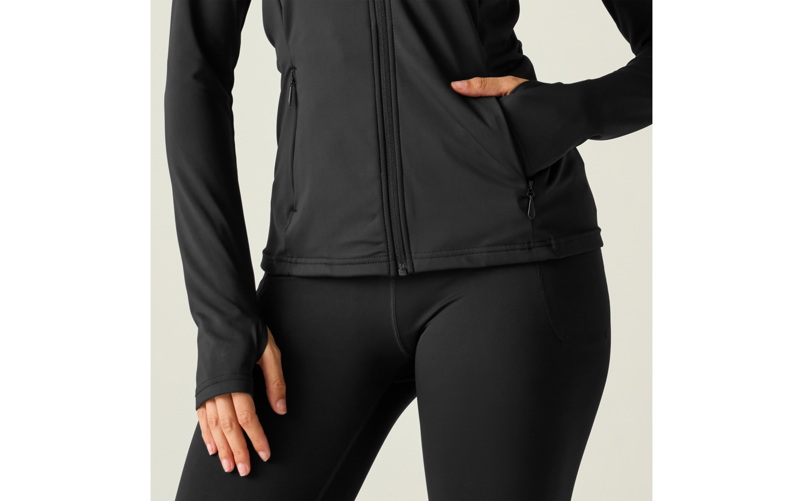 Parte inferior del torso de una persona con chaqueta negra con cremallera y mallas negras, mano derecha en el bolsillo, mano izquierda en el muslo