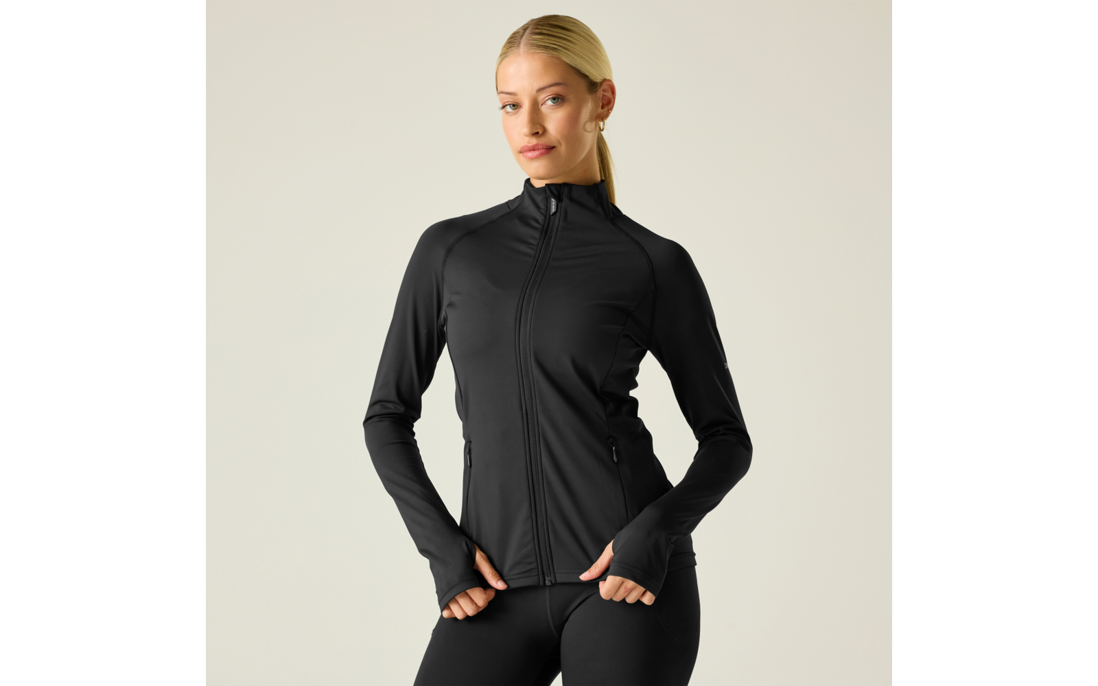 Persona con coleta rubia que lleva una chaqueta deportiva negra ajustada con cremallera y mallas negras, manos en el borde de la chaqueta, fondo liso y claro