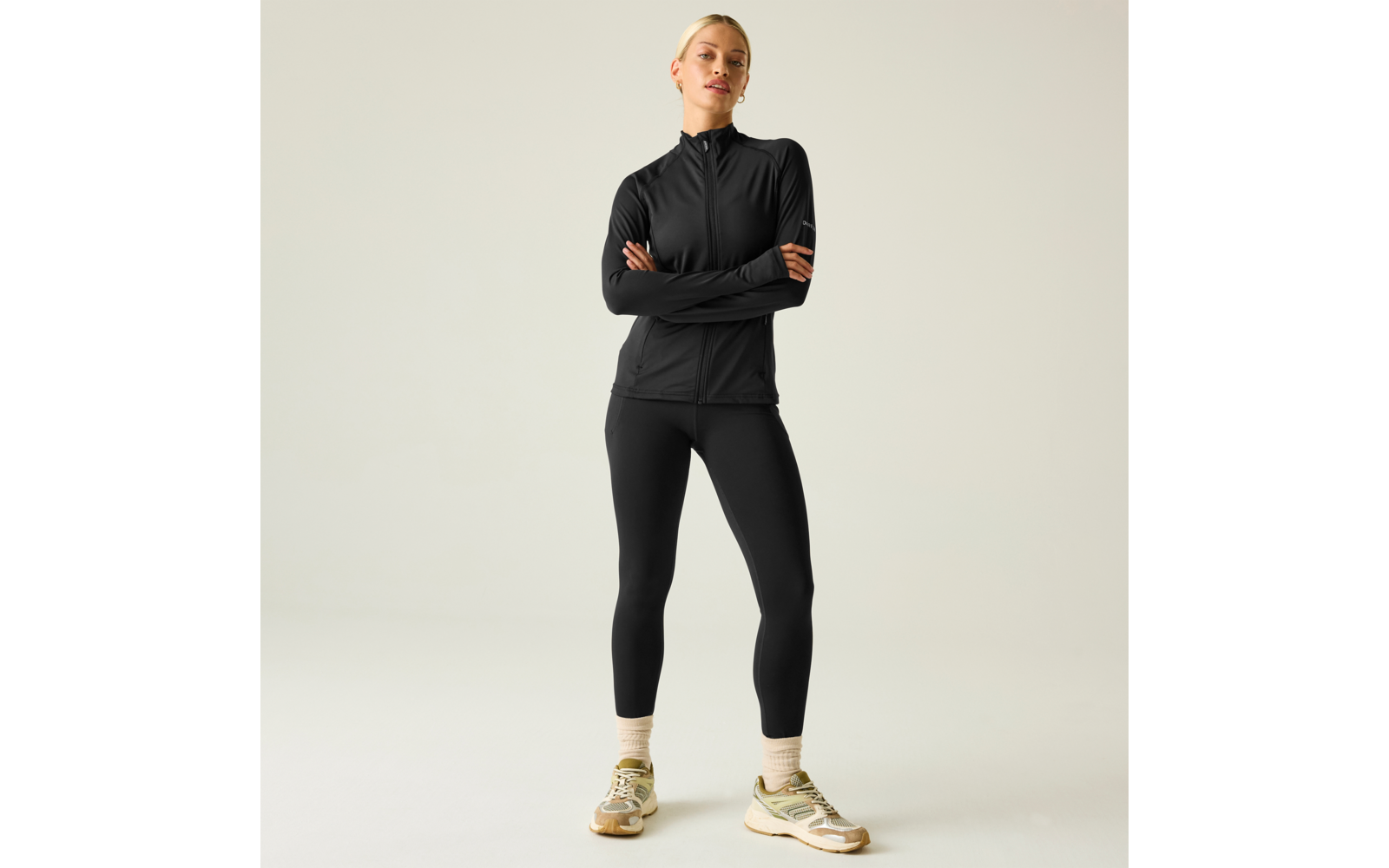 Mujer de cuerpo entero sobre fondo claro, lleva chaqueta negra con cremallera y leggings negros, brazos cruzados, zapatillas beige y verdes