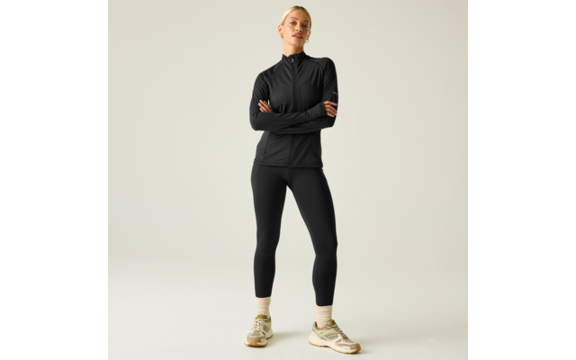 Mujer de cuerpo entero sobre fondo claro, lleva chaqueta negra con cremallera y leggings negros, brazos cruzados, zapatillas beige y verdes