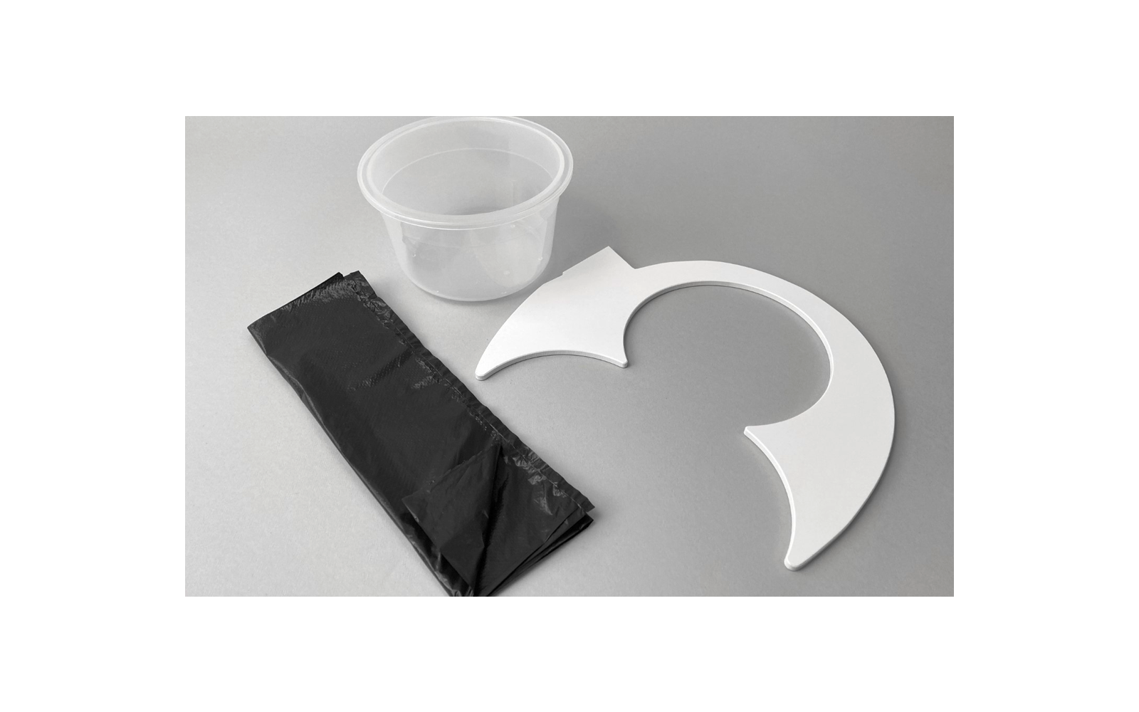 Un gobelet en plastique transparent, un sac-poubelle noir roulé et une pièce en plastique blanche en forme de demi-cercle sur une surface grise
