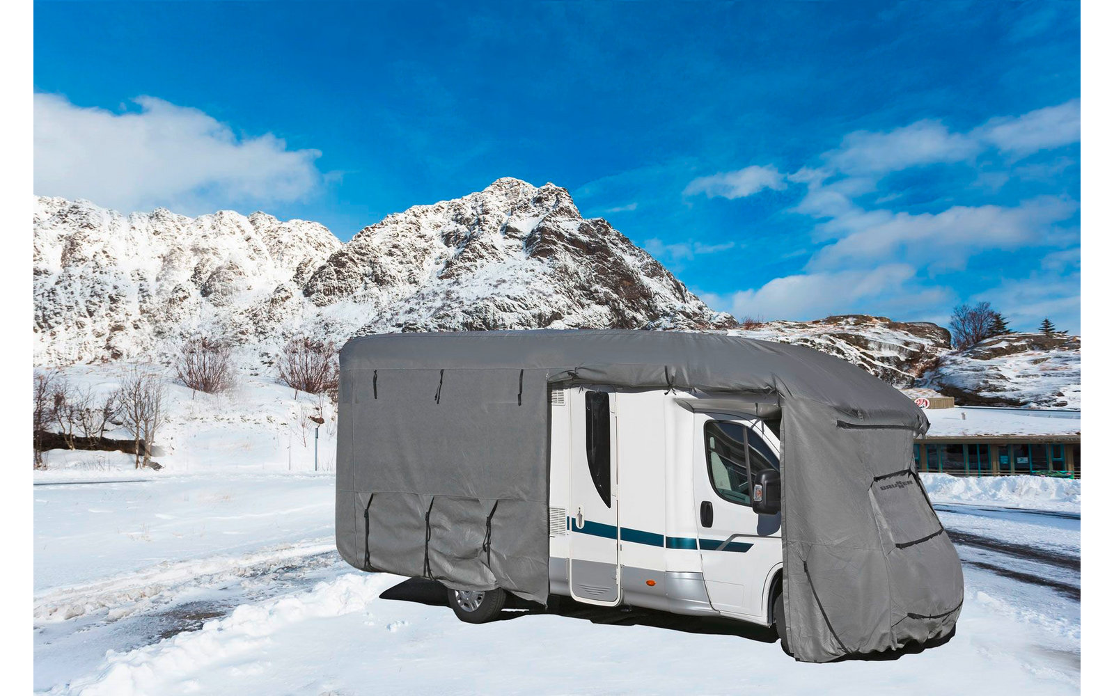 Ein Wohnmobil mit grauer Abdeckung auf schneebedecktem Boden vor schneebedeckten Bergen und blauem Himmel