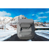 Auto mit grauer Abdeckung auf einer verschneiten, asphaltierten Straße; im Hintergrund schneebedeckte Berge, kahle Bäume, niedrige Gebäude und blauer Himmel mit Wolken