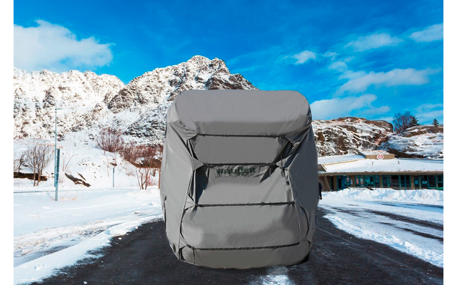 Auto mit grauer Abdeckung auf einer verschneiten, asphaltierten Straße; im Hintergrund schneebedeckte Berge, kahle Bäume, niedrige Gebäude und blauer Himmel mit Wolken