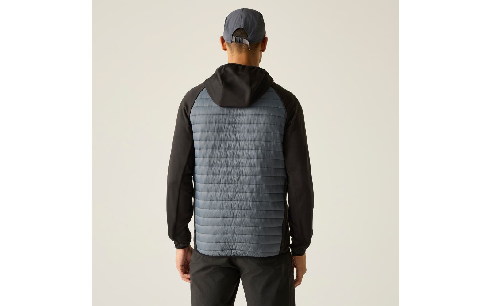 Person von hinten, trägt Jacke mit hellblau-grauem gesteppte(m) Rückenpaneel, schwarze Ärmel und Kapuze, graue Kappe, dunkle Hose, heller Hintergrund