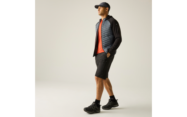 Mann mit Baseballcap, Jacke mit gesteppter Vorderseite und schwarzen Ärmeln, orangefarbenem T-Shirt, schwarzen Shorts, schwarzen Socken und Wanderschuhen, Hände in den Taschen, vor hellem einfarbigem Hintergrund