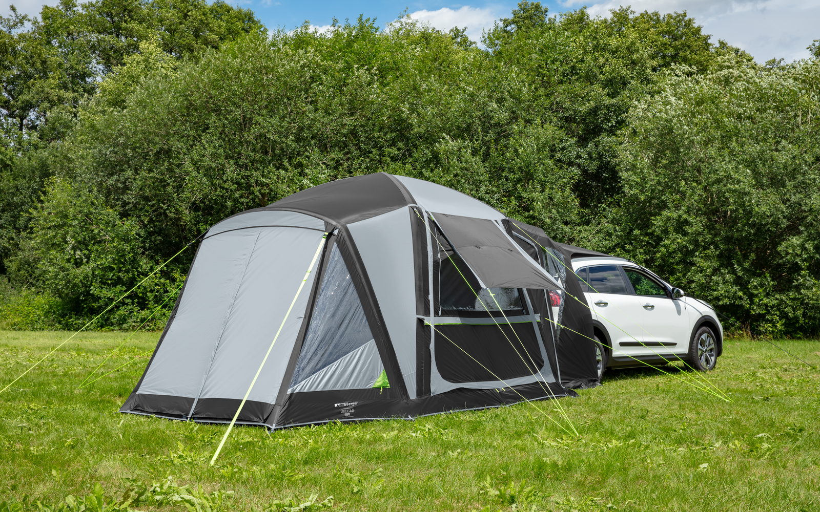 Graues aufgebautes Zelt neben einem weißen Auto auf einer grünen Wiese, im Hintergrund Bäume und wolkiger Himmel