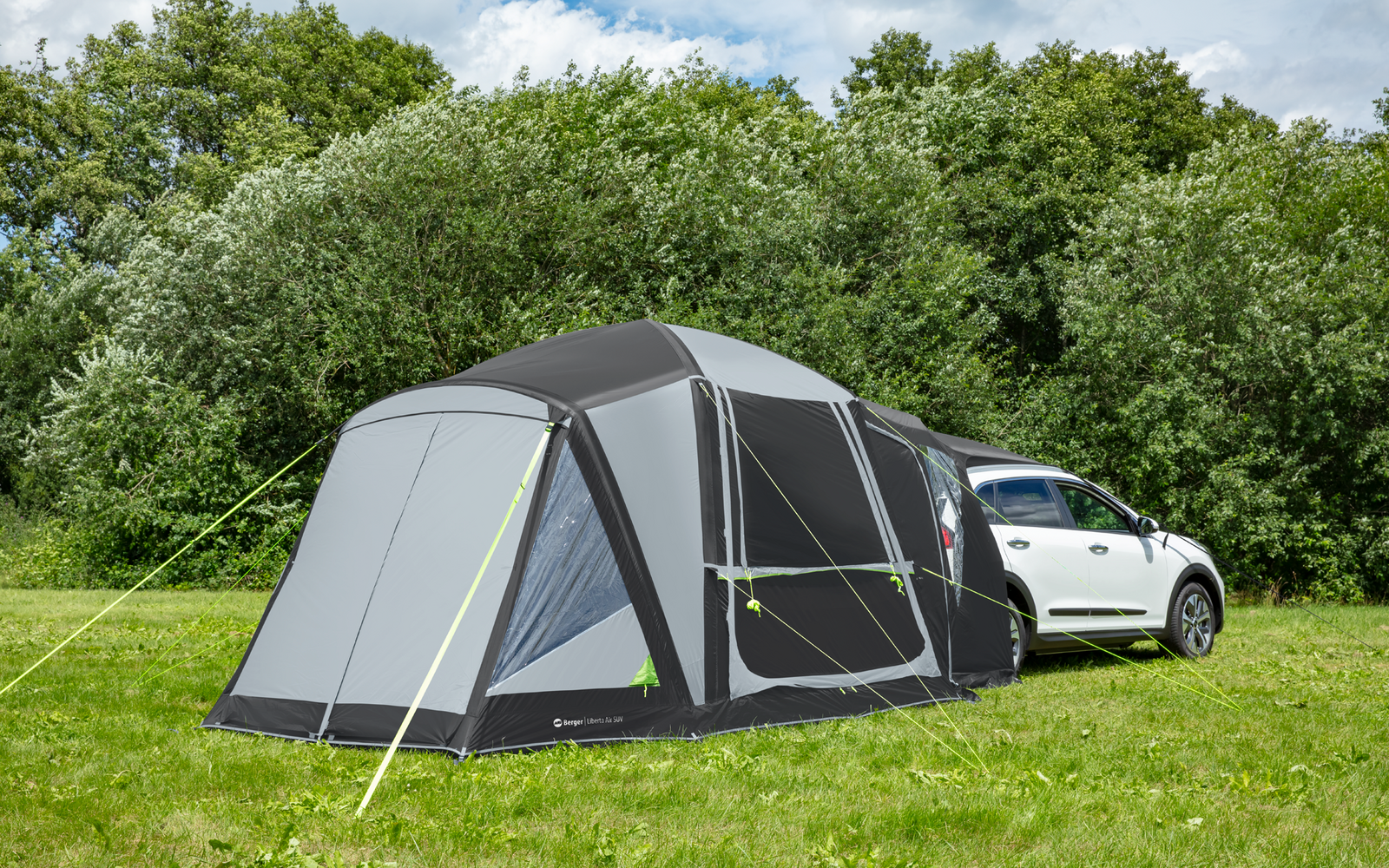 Weißes Auto mit großem dunkelgrauem Vorzelt auf grüner Wiese vor Bäumen und teilweise bewölktem Himmel