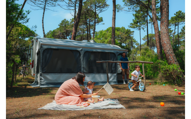 Motorhome with extended awning among pine trees; in front on a blanket a woman and a toddler with toys; to the right a boy on a balance bike and a man next to the camper; colored balls on the grass