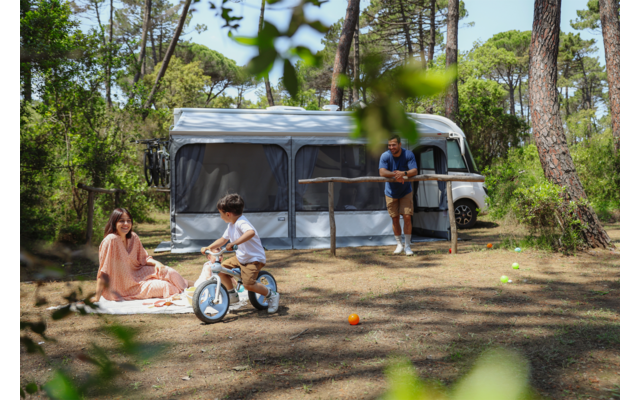 Motorhome with extended awning in a pine-wood clearing; woman sitting on a blanket, child on a balance bike, man leaning against the motorhome; toy balls on the ground