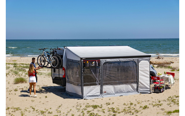Camping vehicle with awning and two bicycles mounted on its rear on a sandy beach, two people standing nearby, sea on the horizon
