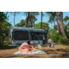 Motorhome with extended awning among pine trees; in front on a blanket a woman and a toddler with toys; to the right a boy on a balance bike and a man next to the camper; colored balls on the grass