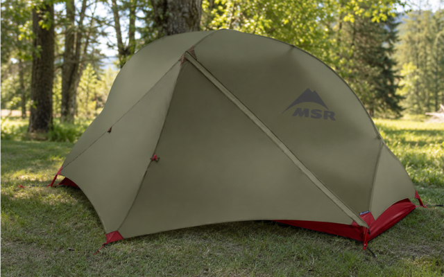 Tente en dôme verte avec logo MSR sur l'herbe, renforts rouges aux angles, arbres et lumière du soleil en arrière-plan
